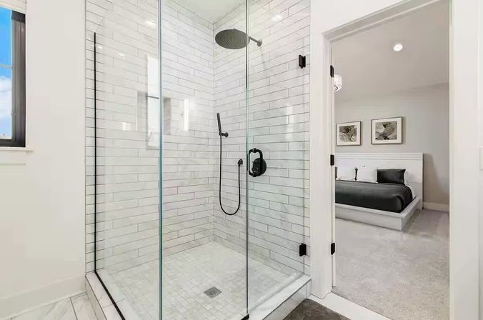 Glass-enclosed walk-in shower with white subway tile and matte-black rain and handheld fixtures, opening to a neutral-toned ensuite bedroom.