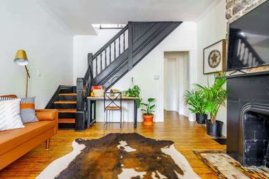 Cozy urban townhouse living room with black painted wooden staircase, cowhide rug on hardwood floors, tan leather sofa with striped pillows, small desk under the stairs, potted green plants, and a black fireplace with wall-mounted TV.