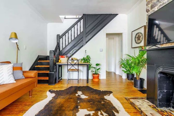 Cozy urban townhouse living room with black painted wooden staircase, cowhide rug on hardwood floors, tan leather sofa with striped pillows, small desk under the stairs, potted green plants, and a black fireplace with wall-mounted TV.