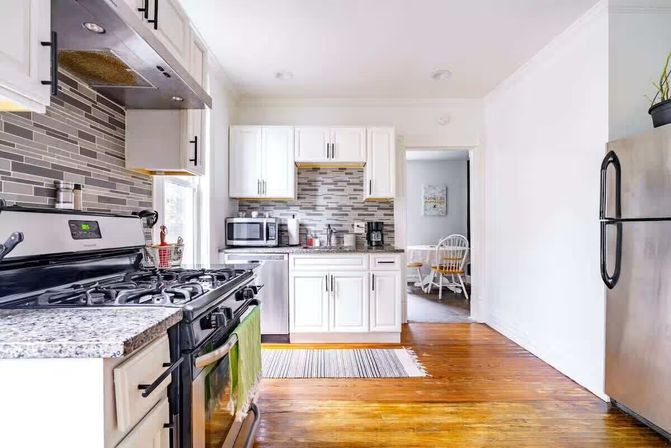 Bright airy modern kitchen with white cabinets, gray mosaic tile backsplash, stainless gas stove and refrigerator, granite countertops, microwave and coffee maker, warm hardwood floors and a small dining nook