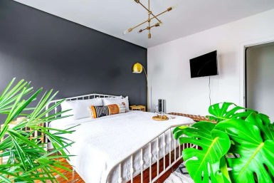 Modern bedroom interior with white metal bed against a charcoal accent wall, gold arc floor lamp, wall-mounted TV, patterned pillows, hardwood floors and large tropical houseplants in foreground.