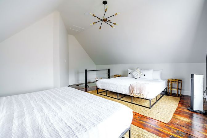 Bright airy loft bedroom with sloped ceiling, two low metal platform beds dressed in white linens, woven jute rug over polished hardwood floors, modern sputnik chandelier and small wooden bedside tables.