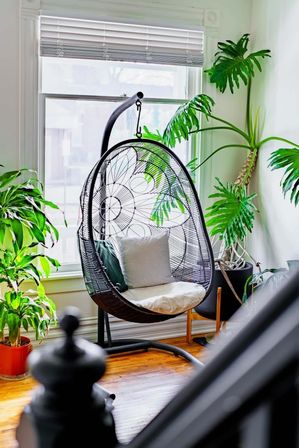 Sunlit boho reading nook with a black woven hanging egg chair on a stand by a large window, striped cushion and tropical houseplants on warm hardwood floors.