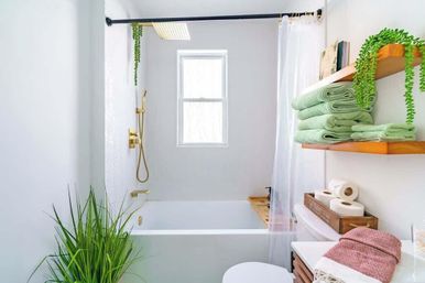 Bright spa-like modern bathroom with white bathtub and gold shower fixtures, window letting in natural light, clear shower curtain, stacked green towels on wooden shelves, potted plant and wooden storage with toilet paper