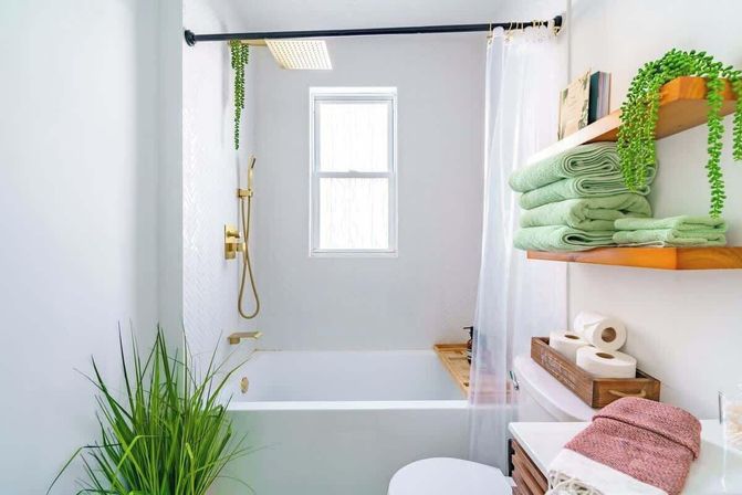 Bright spa-like modern bathroom with white bathtub and gold shower fixtures, window letting in natural light, clear shower curtain, stacked green towels on wooden shelves, potted plant and wooden storage with toilet paper