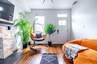 Bright sunlit living room with hardwood floors, orange velvet sofa, hanging rattan egg chair, tall potted plants, exposed brick fireplace and front entry door.