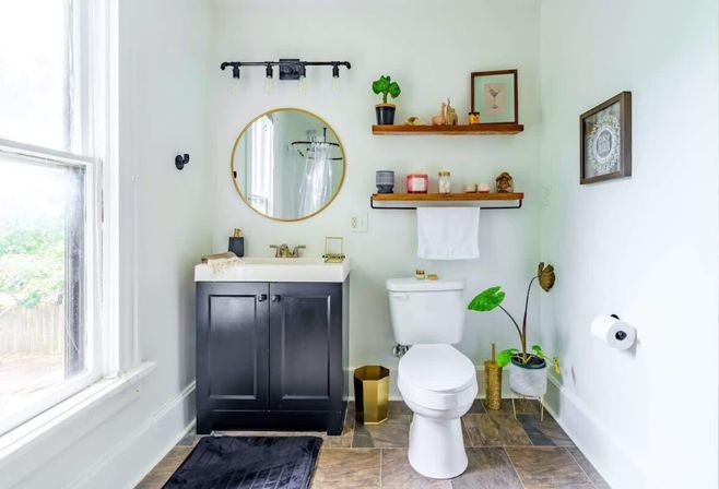 Bright modern bathroom with a black vanity and brass fixtures, round mirror, wooden floating shelves with candles and decor, white toilet, potted green plants, and a large window letting in natural light