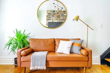 Cozy modern living room interior with tan leather sofa on hardwood floor, round gold mirror above, striped and patterned throw pillows, gray fringed throw, tall green houseplant and brass floor lamp against a white wall.