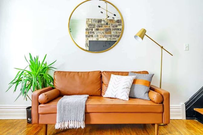 Cozy modern living room interior with tan leather sofa on hardwood floor, round gold mirror above, striped and patterned throw pillows, gray fringed throw, tall green houseplant and brass floor lamp against a white wall.