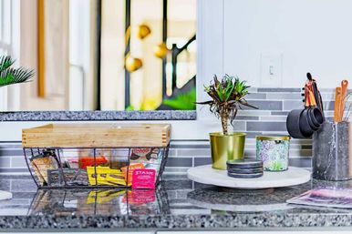 Bright modern kitchen countertop vignette with a wire snack basket, gold potted plant on a marble tray, stacked coasters and hanging measuring cups against a gray subway-tile backsplash.