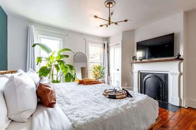 Sunlit cozy bedroom with fluffy white bedding and leather accent pillow, large indoor plants by tall windows, arched floor mirror, wall-mounted TV above a white fireplace, hardwood floors and brass chandelier.