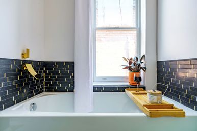 Bright modern bathroom with a white bathtub, black-and-gold subway tile and gold faucet, window, potted plant and bamboo bath tray holding a candle and wine glass.