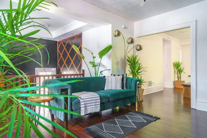 Bright open-plan living room with emerald velvet sofa, patterned throw and rug, brass arc floor lamp, potted indoor plants, geometric wall accent and dark hardwood floors.