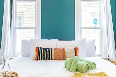 Sunlit bedroom with teal accent wall, two tall windows, white bedding, a textured multicolor lumbar pillow, and three rolled green towels on a plush blanket.