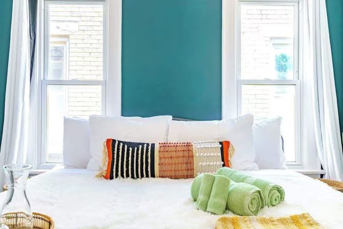 Sunlit bedroom with teal accent wall, two tall windows, white bedding, a textured multicolor lumbar pillow, and three rolled green towels on a plush blanket.