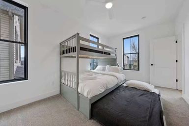 Bright modern bedroom with gray bunk bed and pull-out trundle, crisp white linens, black-framed windows letting in natural light, and neutral carpeted floor.