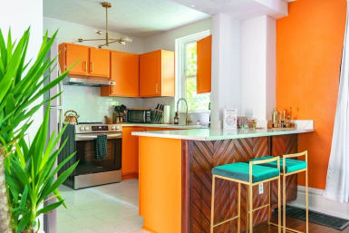 Sunlit modern orange kitchen with white countertops, herringbone wood island front, teal bar stools, stainless steel stove and leafy indoor plant.