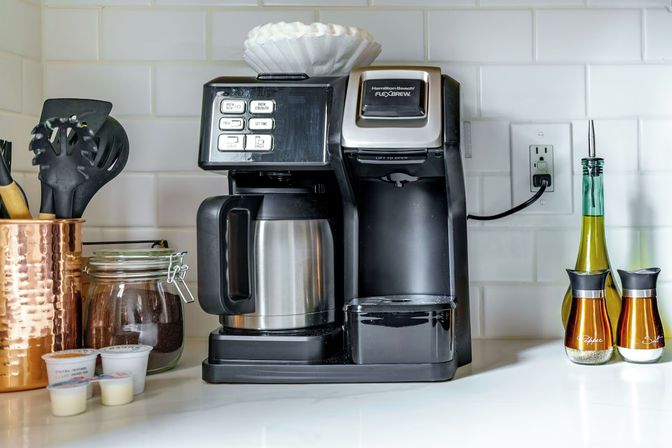 Modern combo single-serve and drip coffee maker on a white subway-tile kitchen counter with stainless carafe, jar of coffee grounds, copper utensil holder, creamer pods, and oil and spice dispensers.