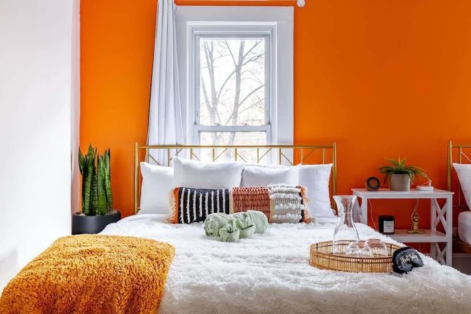 Cozy modern bedroom interior with a bold orange accent wall, gold metal headboard, white fluffy bedding, textured throw pillows and an orange knit throw, indoor plants and a white bedside table beneath a bright window.