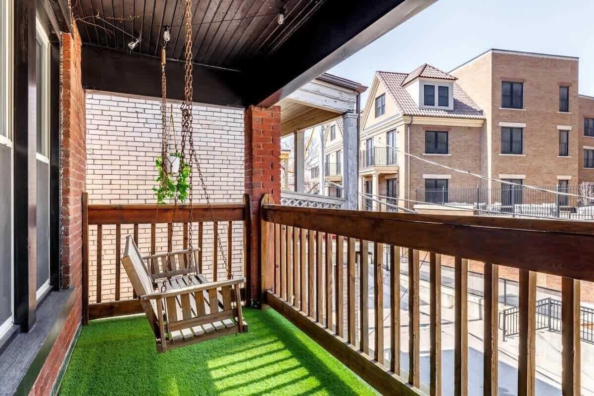 Cozy urban covered balcony with a hanging wooden porch swing, bright faux grass turf, a small hanging plant, and views of neighboring brick townhouses.