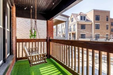Cozy urban covered balcony with a hanging wooden porch swing, bright faux grass turf, a small hanging plant, and views of neighboring brick townhouses.
