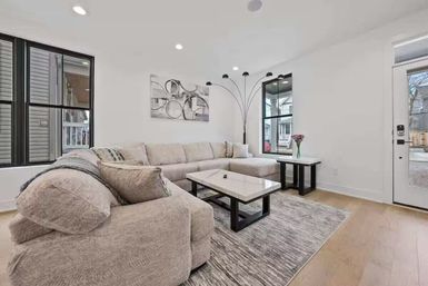 Bright modern living room with cozy light-gray L-shaped sectional, marble-top coffee and side tables, arc floor lamp, large black-trim windows, neutral area rug and light wood floors.