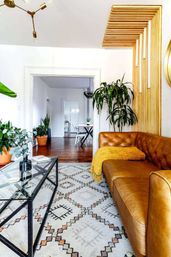 Bright modern boho living room with tan leather tufted sofa and mustard throw, patterned area rug, glass coffee table, indoor plants and wooden slat accent wall opening to a dining area