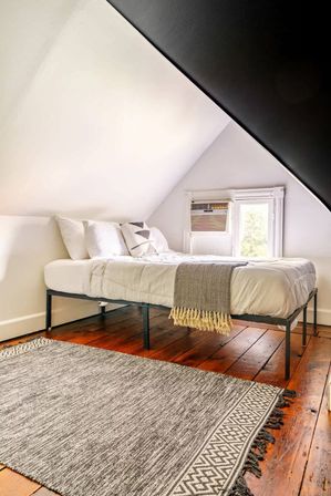 Cozy minimalist attic bedroom with sloped white ceiling, metal-framed bed dressed in white linens and a geometric throw pillow, window air conditioner, warm wood plank floors and a textured gray area rug.