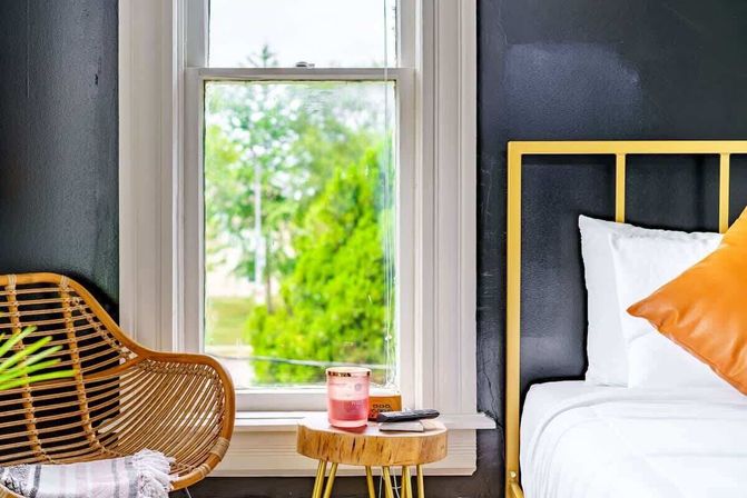 Cozy bedroom corner with a gold metal bedframe, white bedding and orange pillow, rattan chair, small wooden stump side table holding a pink candle and remote, and a window framing green trees outside.