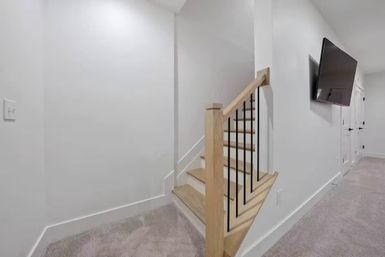 Bright modern basement hallway with light-wood short staircase and black metal balusters, white walls, beige carpeted floor, and wall-mounted flat-screen TV.