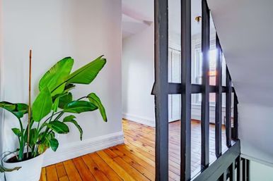 Bright upstairs landing with polished hardwood floors, potted bird-of-paradise plant, white walls and black spindle railing lit by natural light.