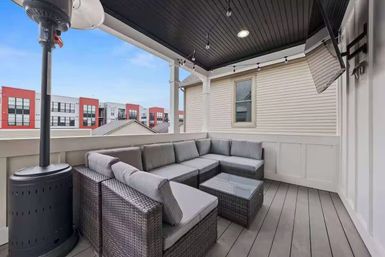 Cozy covered urban rooftop balcony with gray wicker L-shaped sectional, glass-top coffee table, patio heater, wall-mounted TV and string lights overlooking modern apartment buildings.
