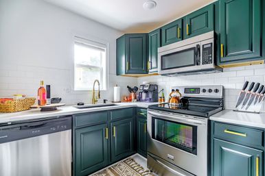 Bright modern kitchen with deep green cabinets and gold hardware, stainless steel stove, microwave and dishwasher, white subway tile backsplash, brass faucet by window, kettle, knife block and coffee station.