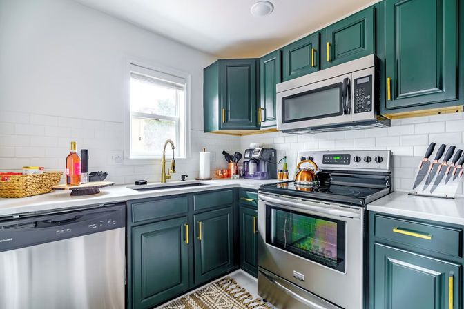Bright modern kitchen with deep green cabinets and gold hardware, stainless steel stove, microwave and dishwasher, white subway tile backsplash, brass faucet by window, kettle, knife block and coffee station.