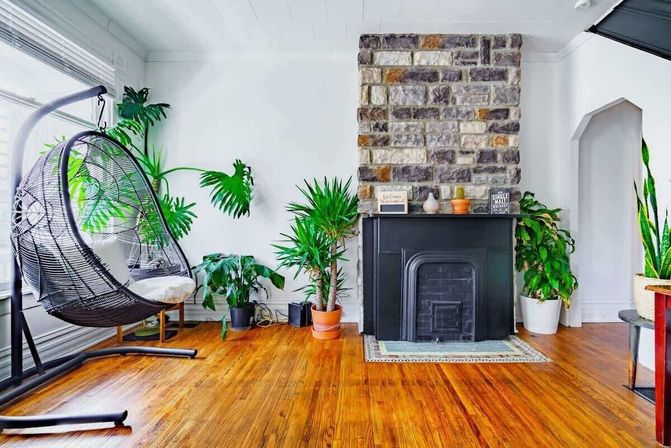 Sunny modern living room with honey hardwood floors, black hanging egg chair by a bright window, assorted potted indoor plants, and a stone chimney with a black fireplace mantel.