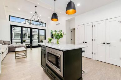 Bright modern open-concept kitchen and living area with marble-topped island and built-in microwave, matte black pendant lights, cozy sectional, large black-framed sliding doors and light hardwood floors.