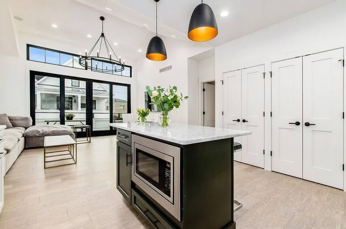 Bright modern open-concept kitchen and living area with marble-topped island and built-in microwave, matte black pendant lights, cozy sectional, large black-framed sliding doors and light hardwood floors.