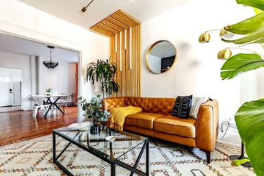 Bright modern living room with a tan tufted leather sofa, glass coffee table, wood-slat accent wall, large indoor plants and patterned boho rug opening to a dining area.