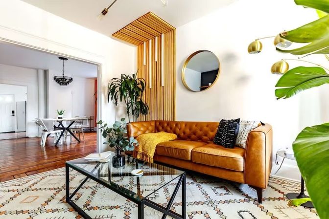 Bright modern living room with a tan tufted leather sofa, glass coffee table, wood-slat accent wall, large indoor plants and patterned boho rug opening to a dining area.