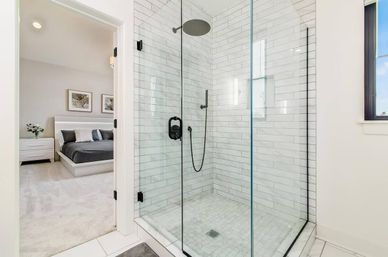 Bright modern ensuite with glass-enclosed corner shower clad in white subway tile, black matte rain showerhead and handheld, and tiled floor drain; open doorway reveals a neutral master bedroom with platform bed and nightstand.