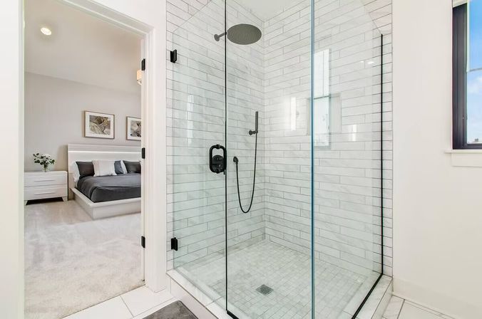 Bright modern ensuite with glass-enclosed corner shower clad in white subway tile, black matte rain showerhead and handheld, and tiled floor drain; open doorway reveals a neutral master bedroom with platform bed and nightstand.