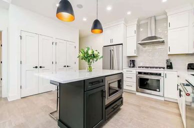 Bright modern kitchen with a dark central island, white shaker cabinets, stainless steel refrigerator and range, built-in microwave, black pendant lights, marble countertops, vase of green leaves, and wood floors.