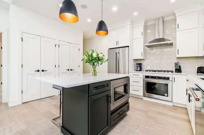Bright modern kitchen with a dark central island, white shaker cabinets, stainless steel refrigerator and range, built-in microwave, black pendant lights, marble countertops, vase of green leaves, and wood floors.