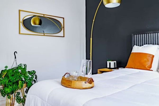 Contemporary bedroom interior with white comforter and black accent wall, gold arched floor lamp, tan leather pillow, potted monstera, and wooden tray with glassware on the bed.