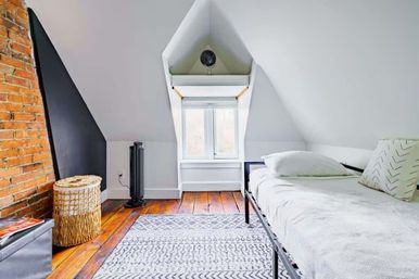 Cozy bright attic bedroom with sloped white ceiling, exposed brick accent wall, single metal bed, hardwood floors, patterned rug, wicker basket and dormer window.
