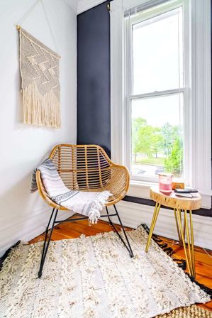 Sunlit boho reading nook with rattan chair and striped throw, macramé wall hanging, textured cream rug, round wood side table and window view of green trees.