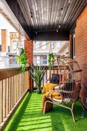 Cozy urban balcony oasis with bright green artificial turf, wicker egg chair draped in a mustard throw, hanging planters, potted snake plant, string lights and red brick facade