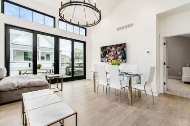 Bright modern open-plan living and dining room in a contemporary apartment with high ceilings and circular chandelier, glass dining table with white chairs and tulips, wall-mounted TV, light hardwood floors, marble-top nesting tables, and large black-framed sliding doors opening to a balcony.
