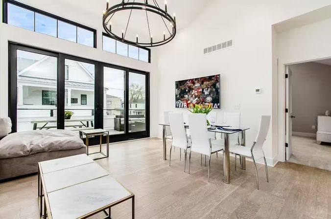 Bright modern open-plan living and dining room in a contemporary apartment with high ceilings and circular chandelier, glass dining table with white chairs and tulips, wall-mounted TV, light hardwood floors, marble-top nesting tables, and large black-framed sliding doors opening to a balcony.