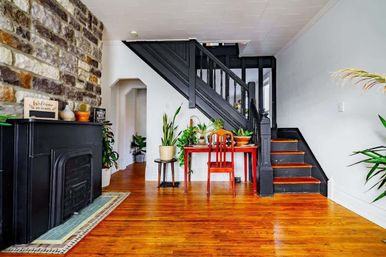 Cozy modern-vintage foyer with glossy hardwood floors, stone accent wall, black vintage fireplace and painted black staircase, red wooden desk and chair topped with potted houseplants